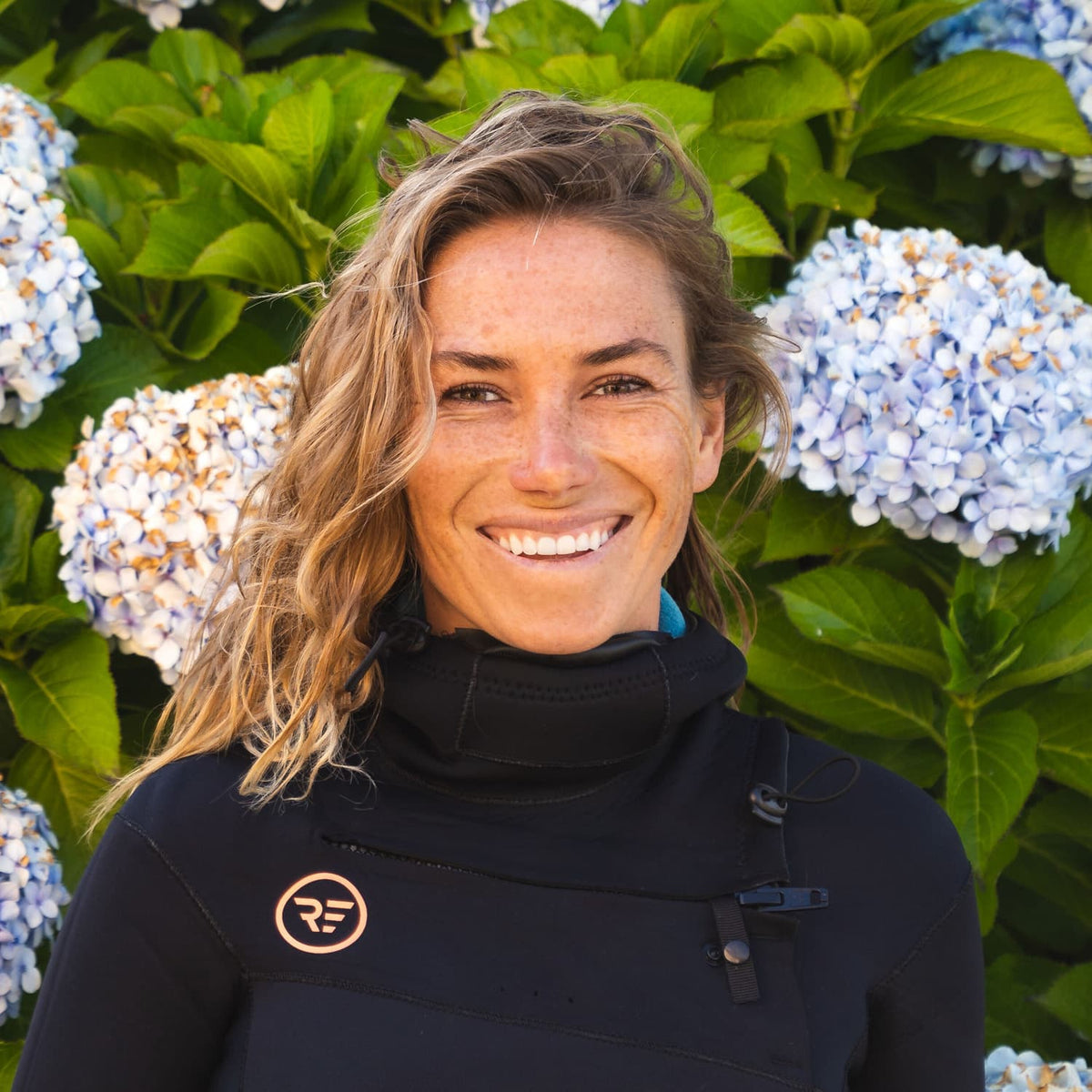 Smiling woman with wavy blonde hair wearing a black high-neck wetsuit standing outdoors in front of green leaves and light purple hydrangea flowers.