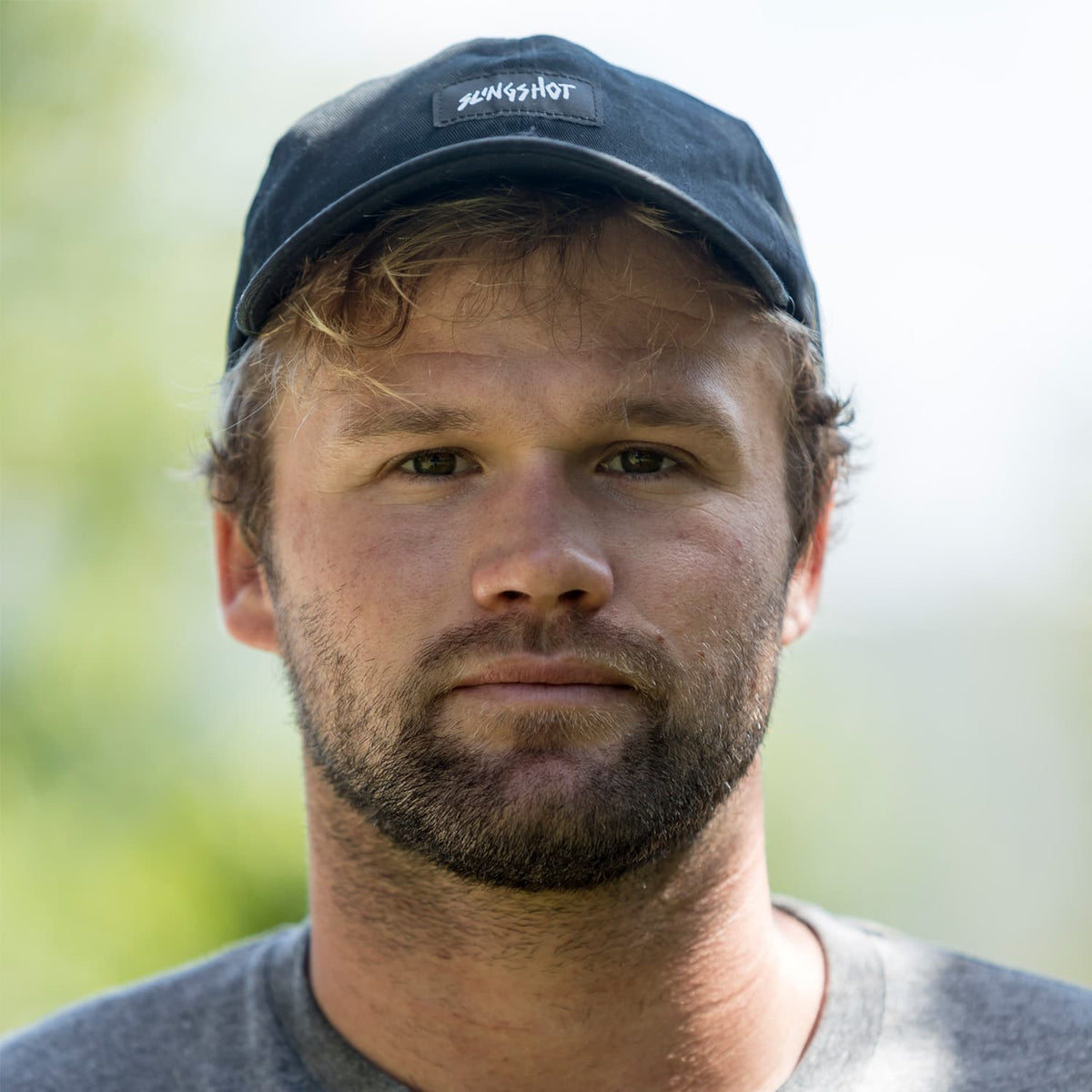 Close-up portrait of a man wearing a black cap with the Slingshot logo, outdoors with a blurred natural green background, showcasing a neutral expression and light beard.