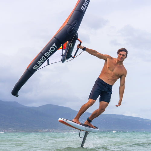 Athlete riding a foil board on the water using a wing with a sleek black and orange design, showcasing the Carbon Uni Handle in action against a mountainous backdrop.