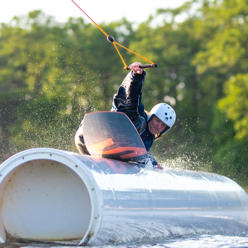 Athlete wearing a helmet and wetsuit riding a 2024 Space Rover foil board on water, gripping a tow handle with water splashing around and green trees in the background.