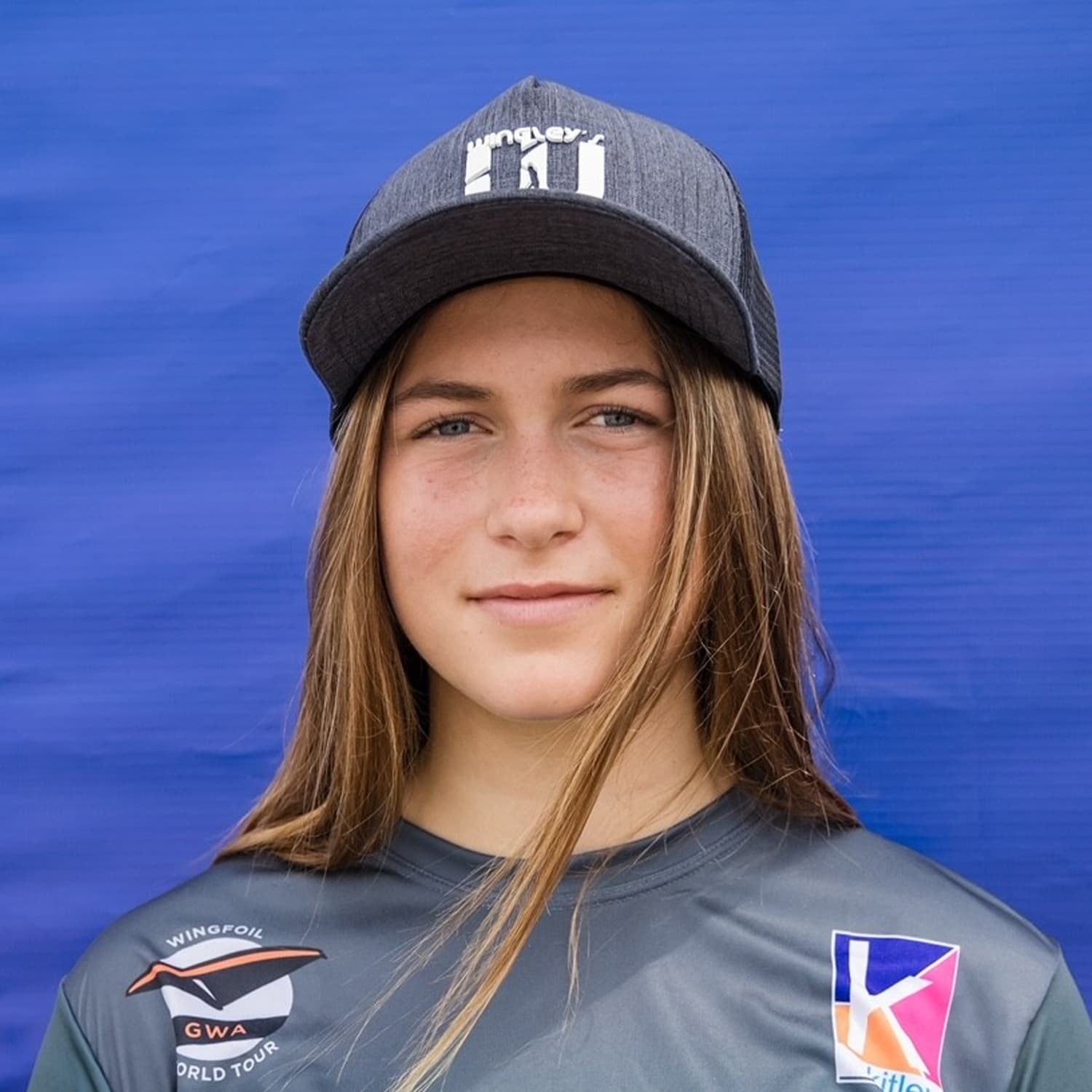 Close-up portrait of a young female athlete wearing a grey cap and a sports jersey with Wingfoil World Tour and kit logos, set against a solid blue background.