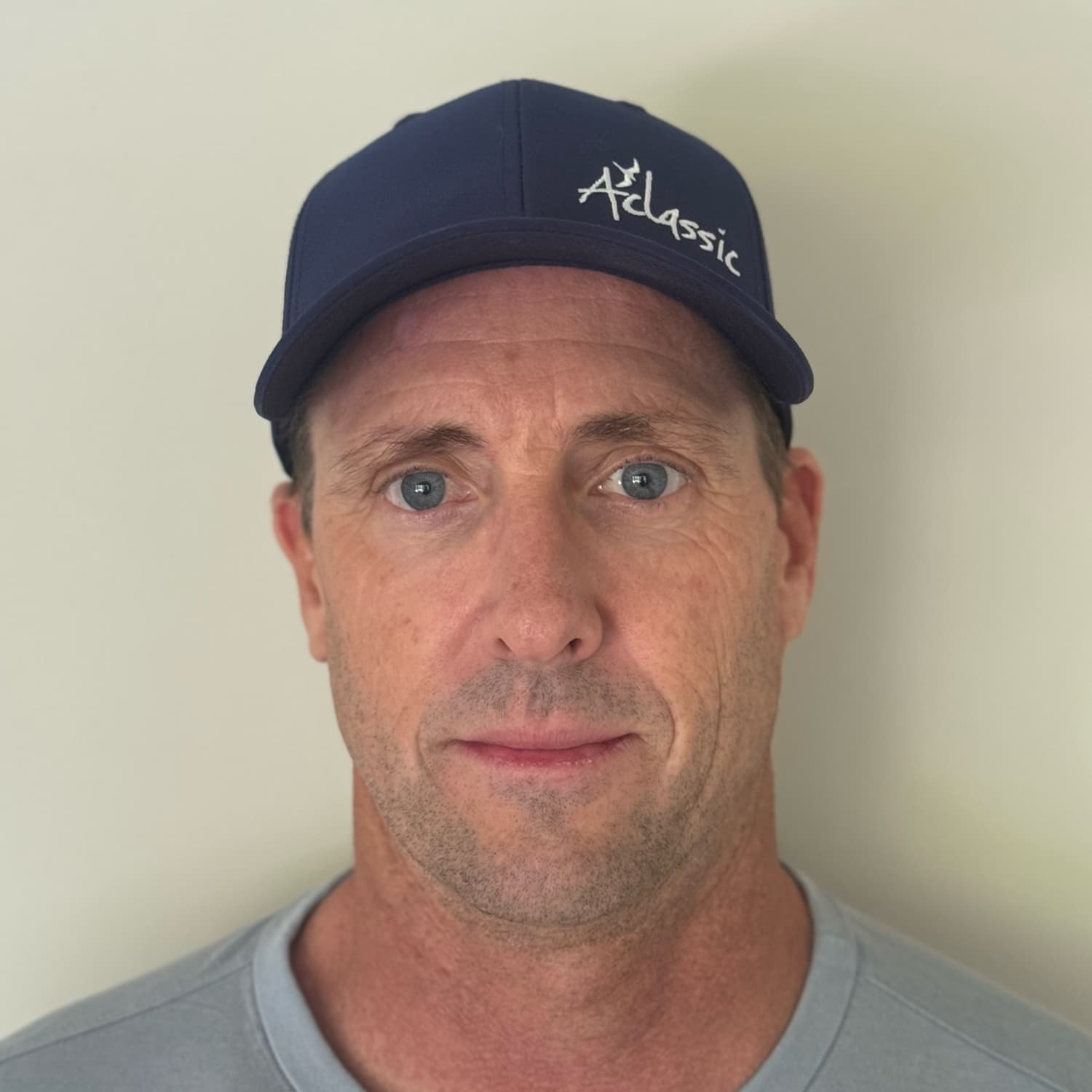 Close-up portrait of a middle-aged man with blue eyes wearing a navy blue cap with "A Classic" embroidered on it, against a plain light background.