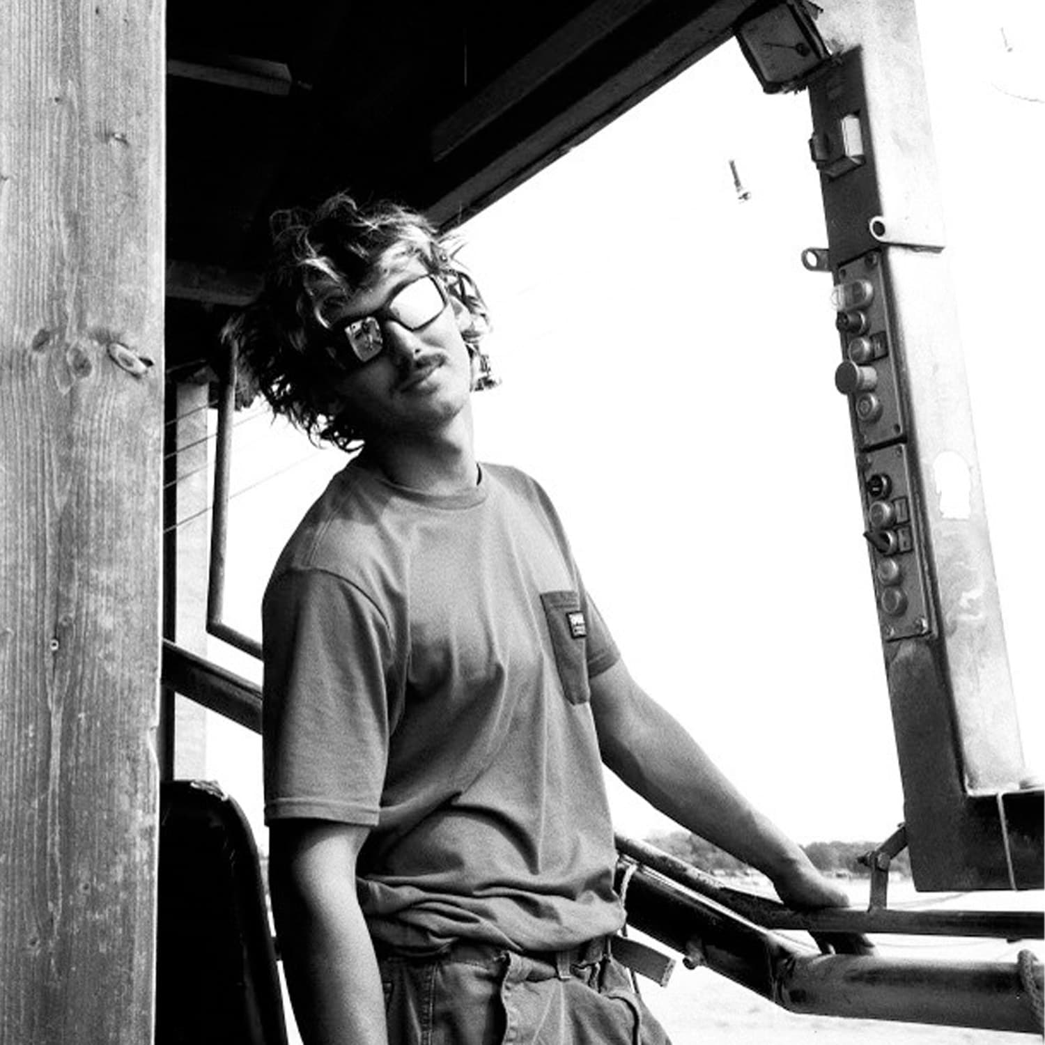 Black and white photo of a young person with curly hair wearing sunglasses and a t-shirt, leaning casually against a wooden beam and a metal structure in an outdoor setting.