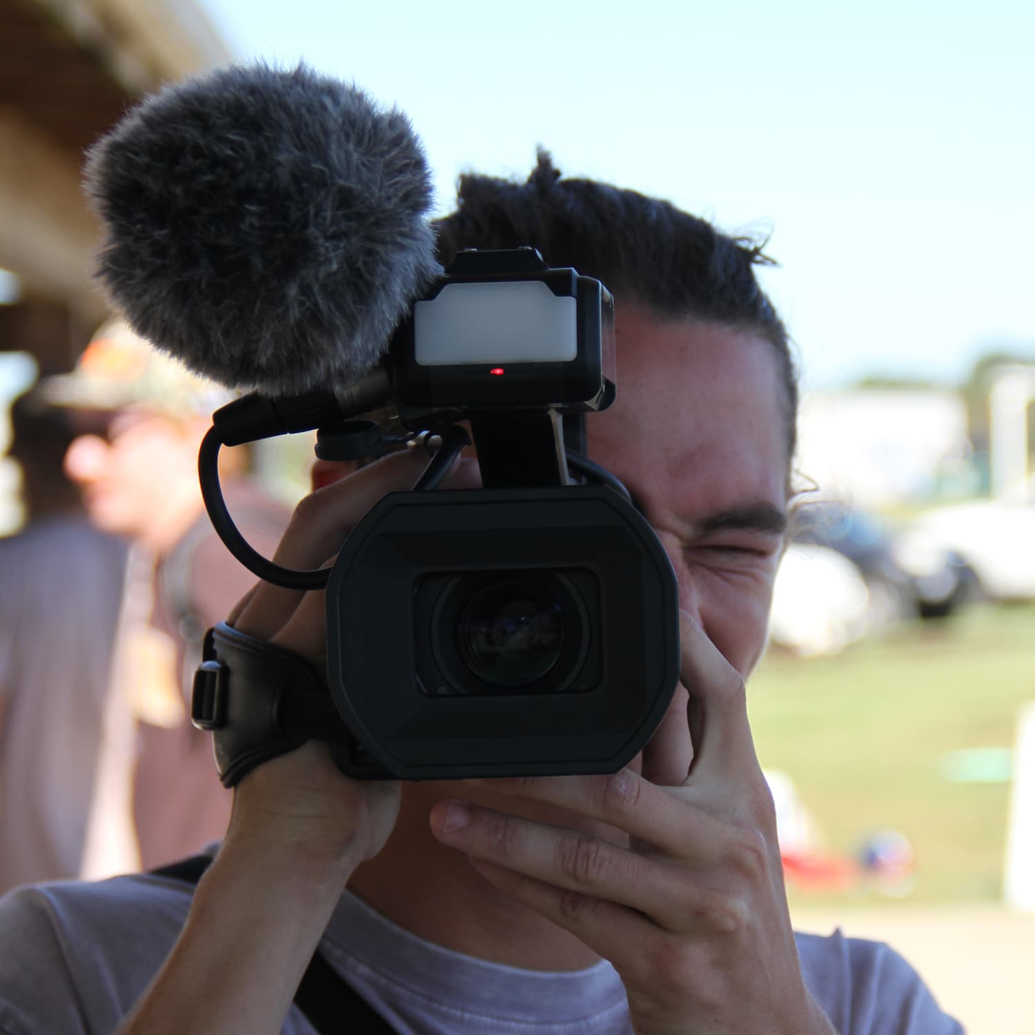 Person holding a professional video camera with a large microphone attachment, filming outdoors on a sunny day with blurred background elements.