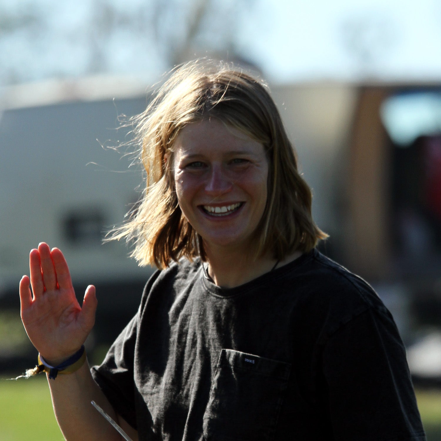 Smiling young person with shoulder-length hair wearing a dark shirt, waving at the camera outdoors with blurred background elements.
