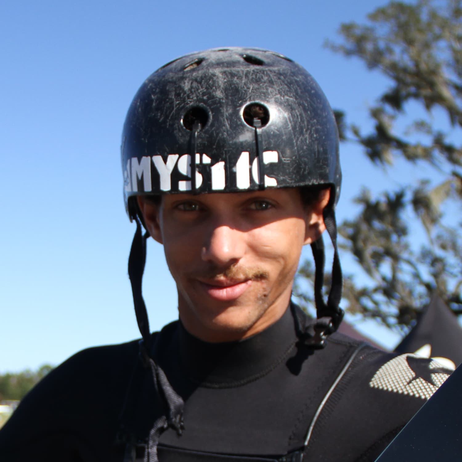 Close-up of a young male athlete wearing a black helmet with visible wear and a black wetsuit, standing outdoors with clear blue sky and trees in the background.