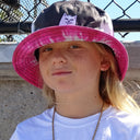 Close-up of a young person with long blonde hair wearing a black and pink tie-dye bucket hat featuring a white cat graphic, standing outdoors in front of a chain-link fence.