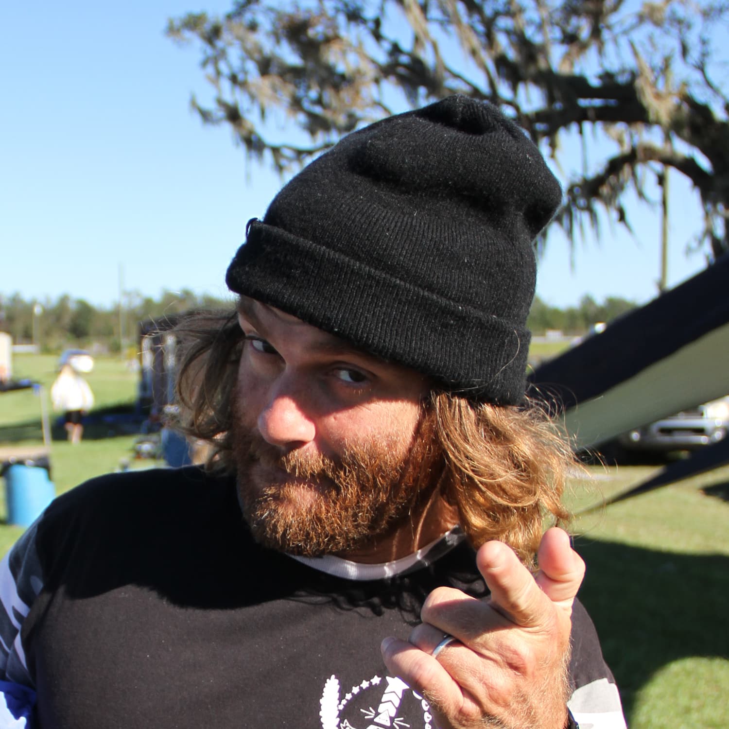 Man wearing a black beanie and dark shirt with a beard and long hair points towards the camera outdoors on a sunny day with trees and people in the background.