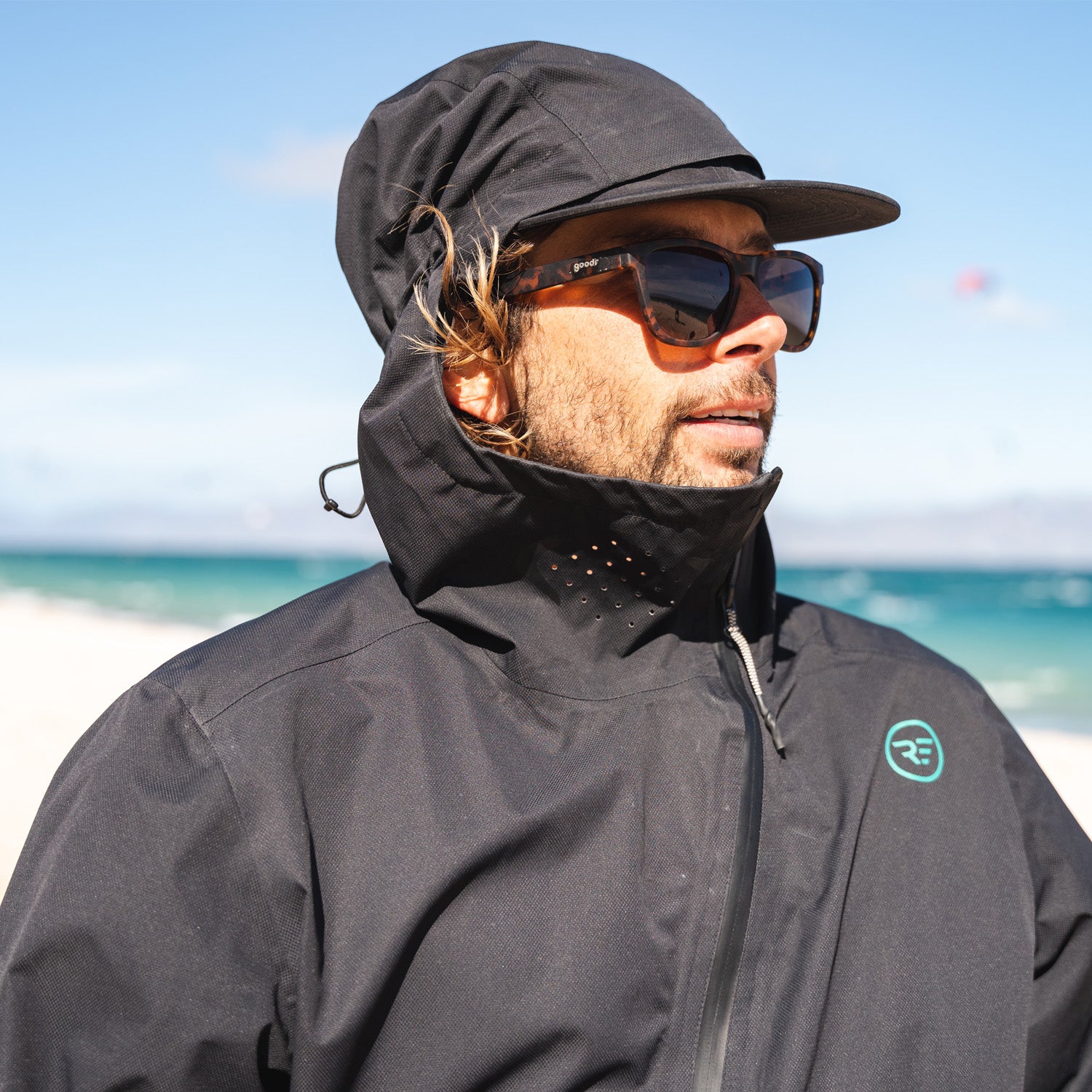 Man wearing a black hooded jacket and cap with sunglasses, standing outdoors on a sunny day near the ocean with gentle waves and a clear sky.