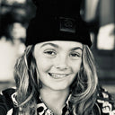 Close-up of a smiling young person wearing a black beanie and patterned shirt, with long hair and a natural outdoor background blurred behind them.
