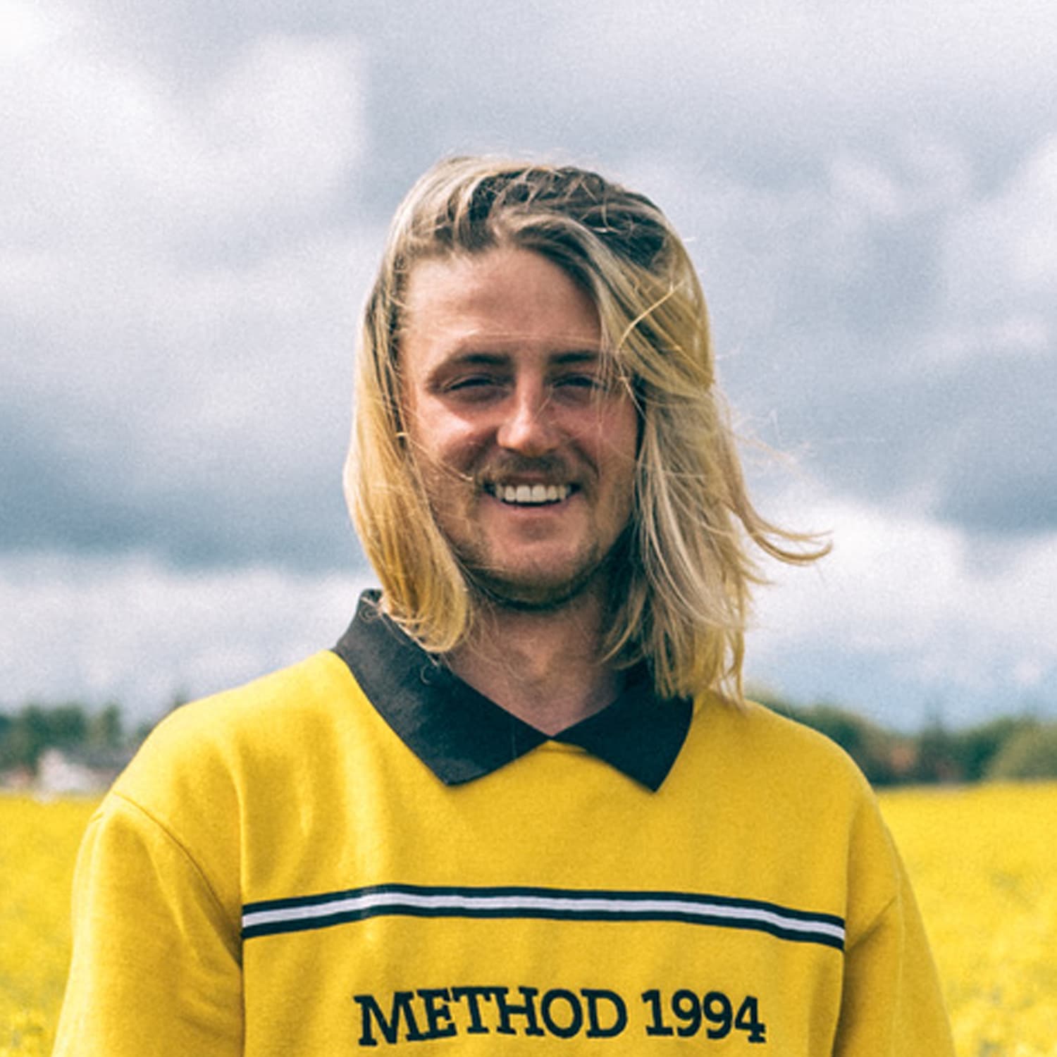 Young man with shoulder-length blonde hair wearing a yellow collared shirt with "METHOD 1994" text, standing outdoors with a cloudy sky and field in the background.