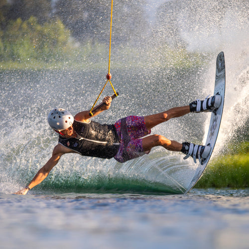 Athlete wearing helmet and life vest riding a 2025 Solo wakeboard, cutting sharply across water while holding a tow rope, creating a large spray of water in bright sunlight.