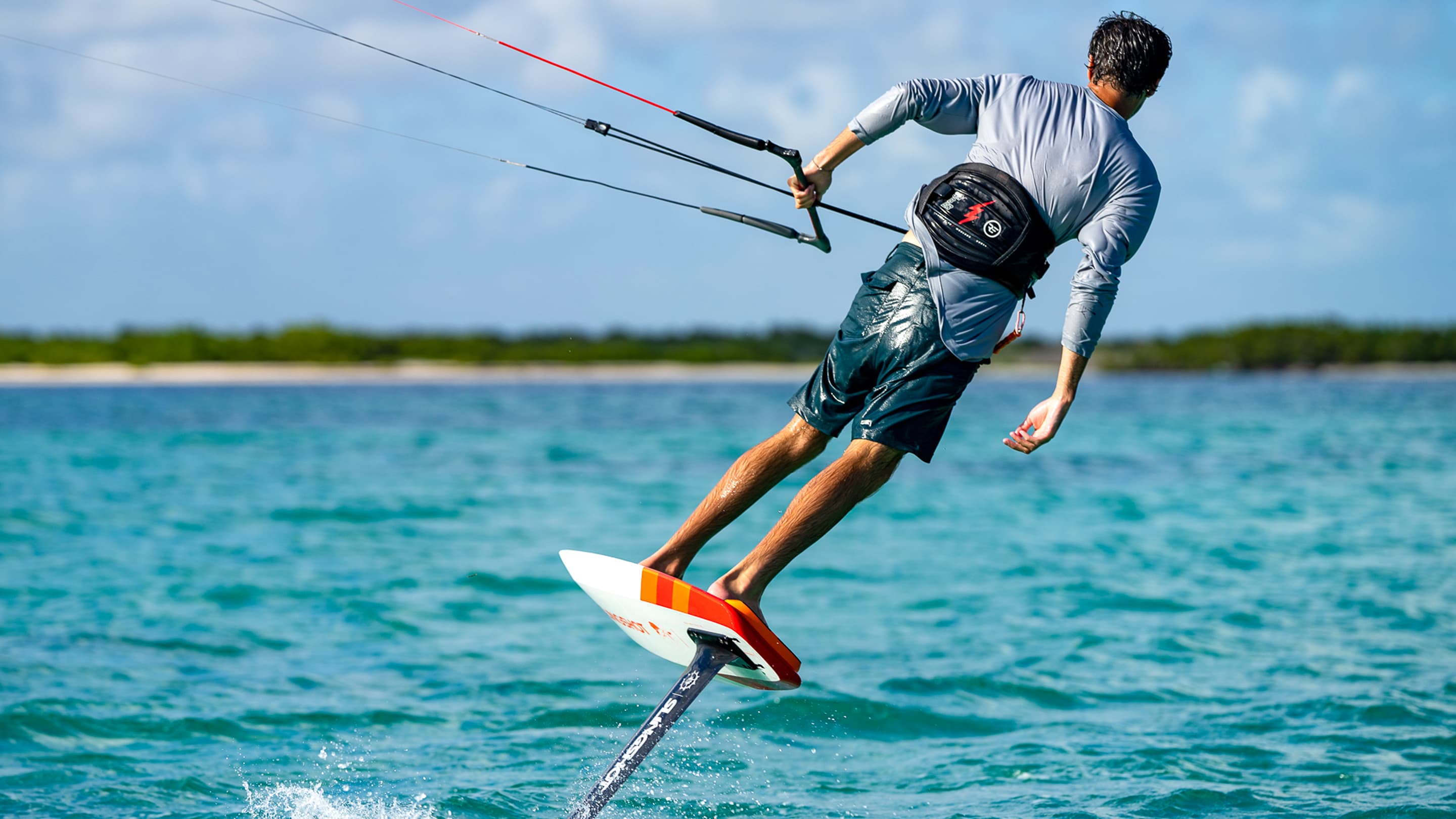 Athlete riding a hydrofoil kiteboard on clear blue water, wearing a harness and controlling the kite with a bar against a backdrop of sky and distant shoreline.