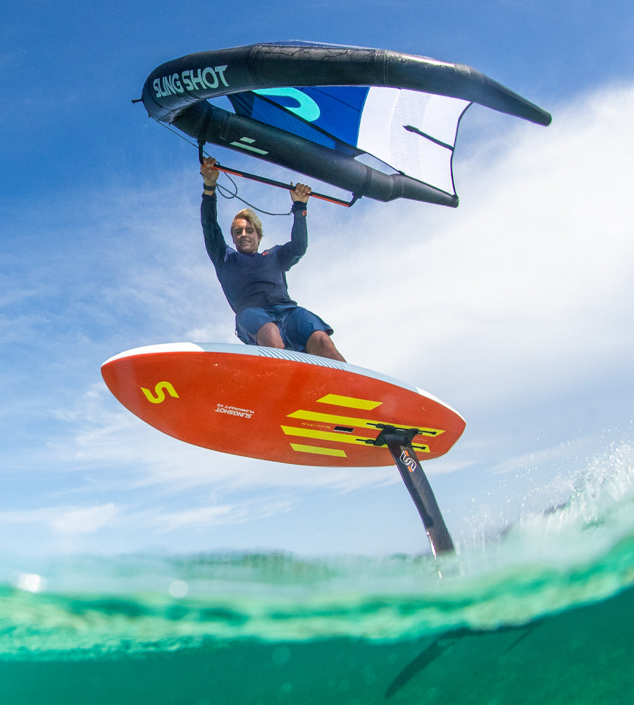 Person holding a Slingshot wing foil above clear turquoise water while riding an orange hydrofoil board under a bright blue sky.