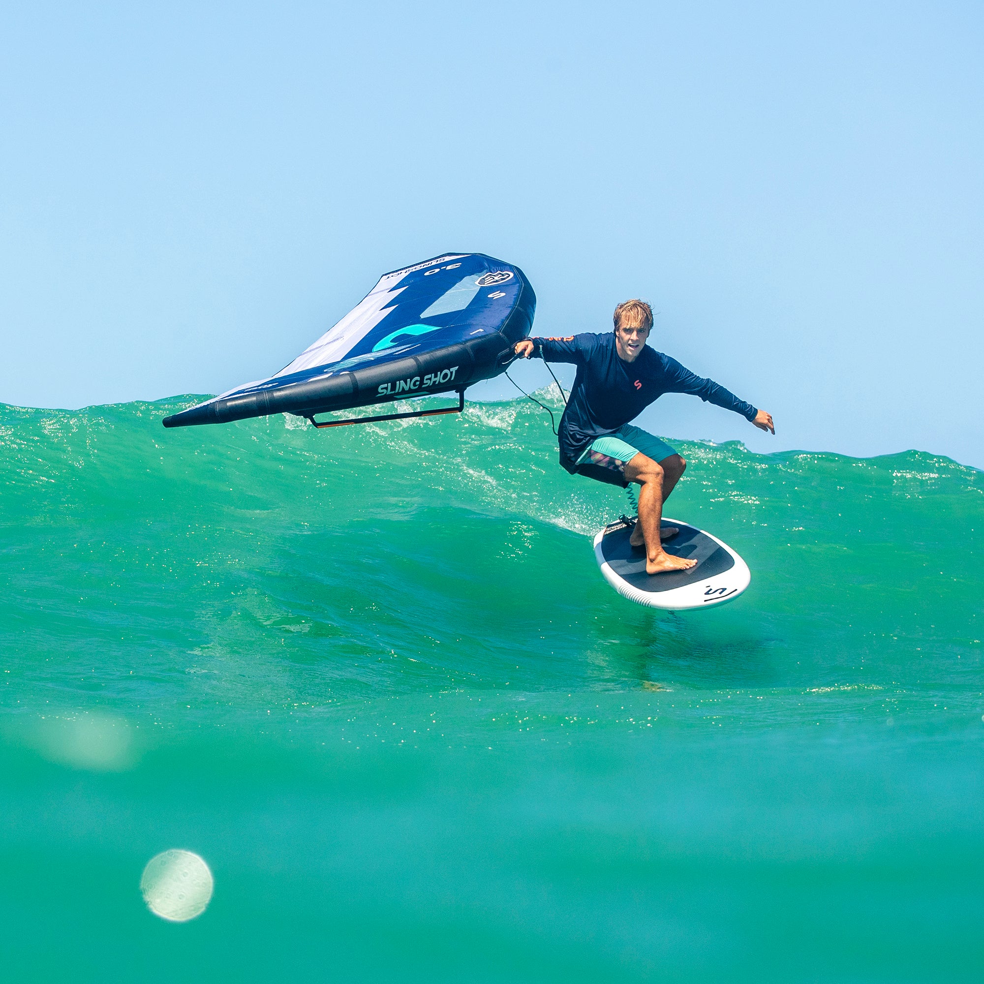 Athlete riding a foil board over a green wave, using the One-Lock Glide Front Wing for efficient glide and controlled handling in everyday water conditions.
