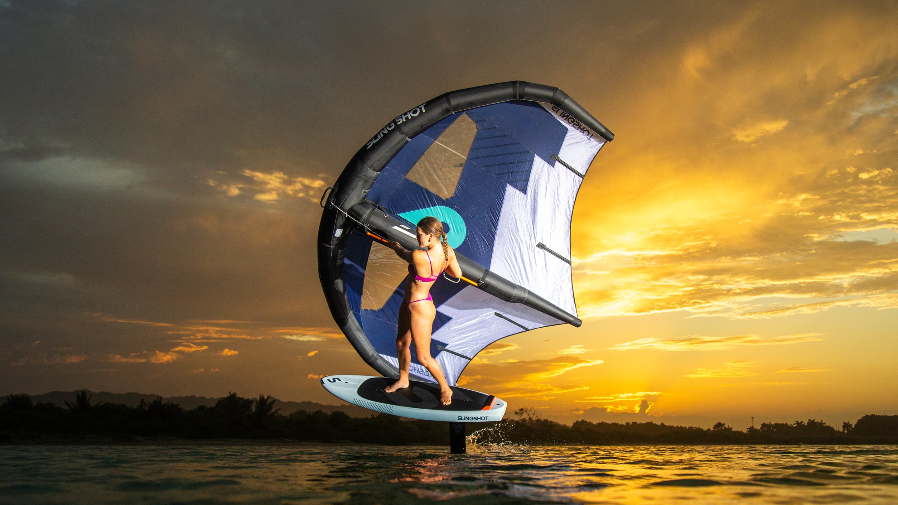 Woman riding a wing foil board on calm water during a vibrant sunset, holding a large inflatable wing with a sleek design and standing on a hydrofoil board.