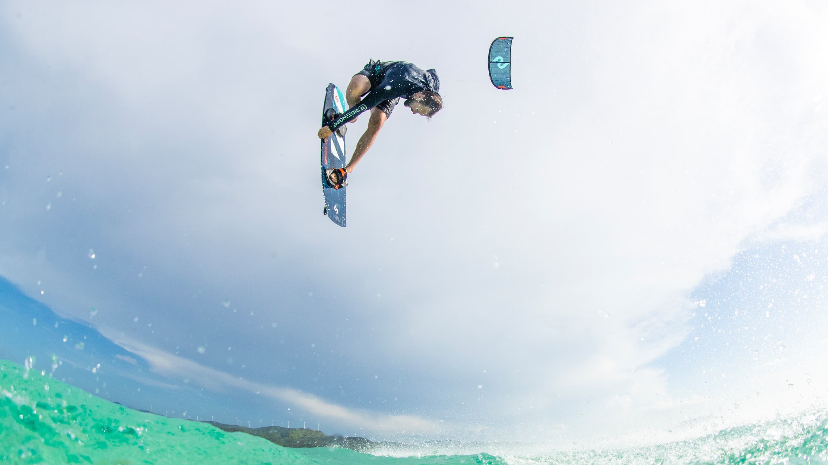 Kiteboarder performing an aerial trick above turquoise water with a cloudy sky backdrop, showcasing dynamic movement and skill in watersports.