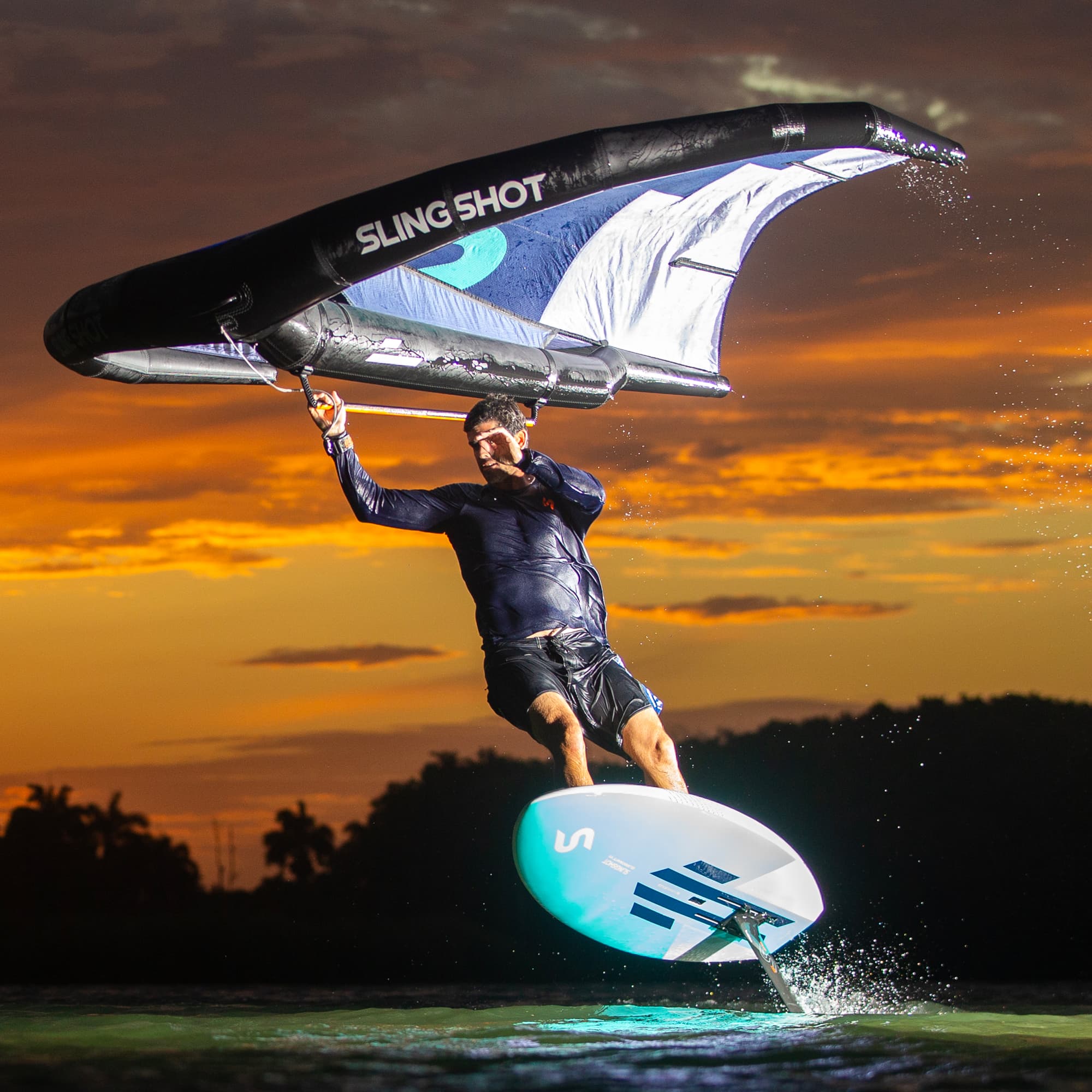 Man riding a foil board on water at sunset, holding the SlingWing V6 wing with Rider Driven Design, showcasing smooth power and stability in wing foiling.