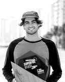 Young man wearing a cap and long-sleeve shirt smiling while holding a hydrofoil board with city buildings blurred in the background.
